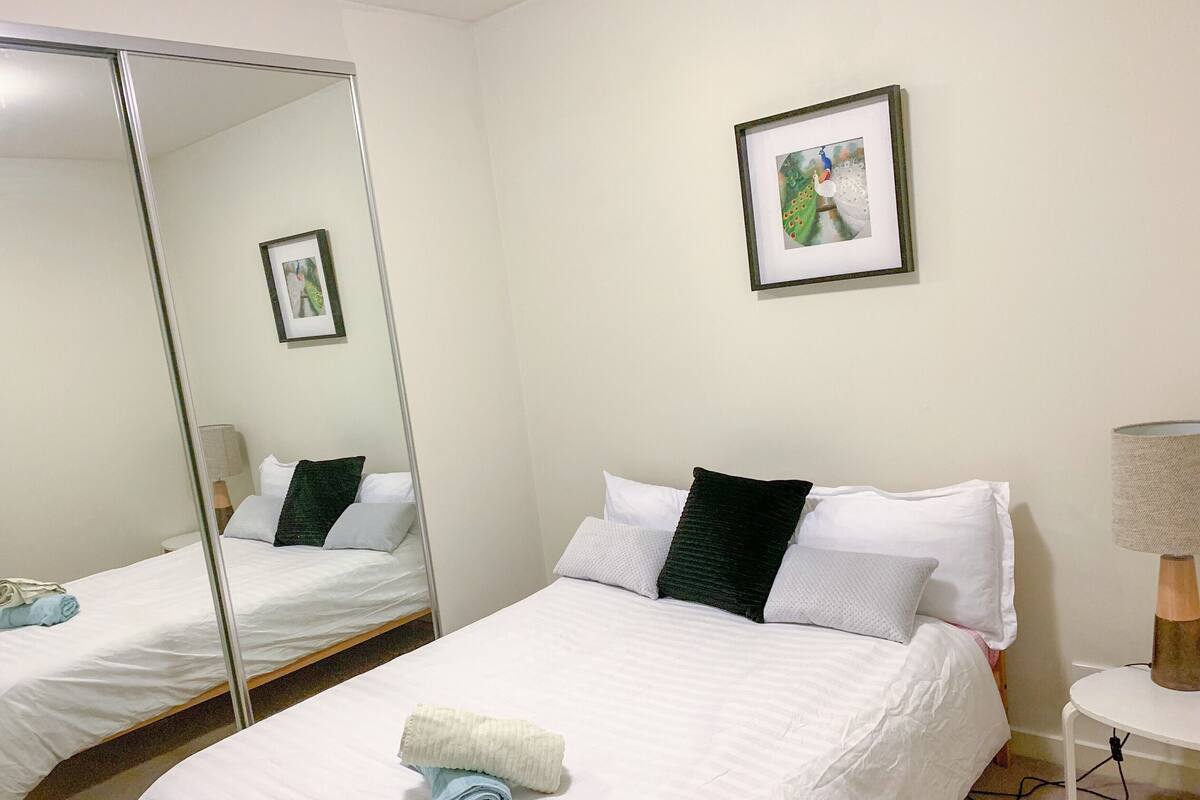 A hotel room with two beds, a large mirror, a framed picture on the wall, and a bedside table with a lamp.