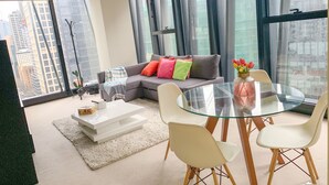 A modern living room with a glass dining table, white chairs, a grey sofa with colorful cushions, and a view of tall buildings through large windows. - Sunshine Boutique Apartments Collins (Melbourne)