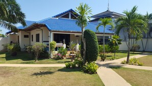 A two-story house with a blue tiled roof, a front porch with a bench, and a well-maintained garden with various plants and a palm tree.