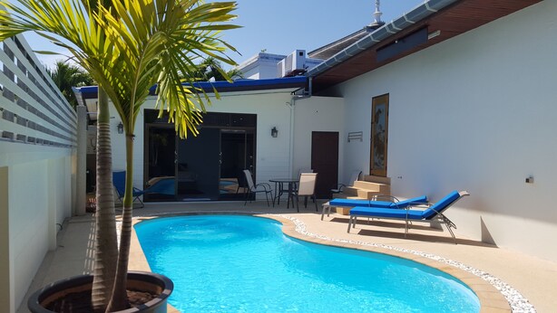 A swimming pool with lounge chairs and a palm tree, surrounded by a white building with a blue roof and a door leading to a covered area.