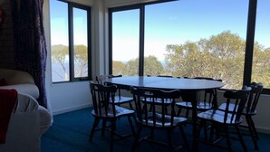 Um quarto com uma mesa redonda e seis cadeiras, janelas amplas com vista para árvores e um céu claro.