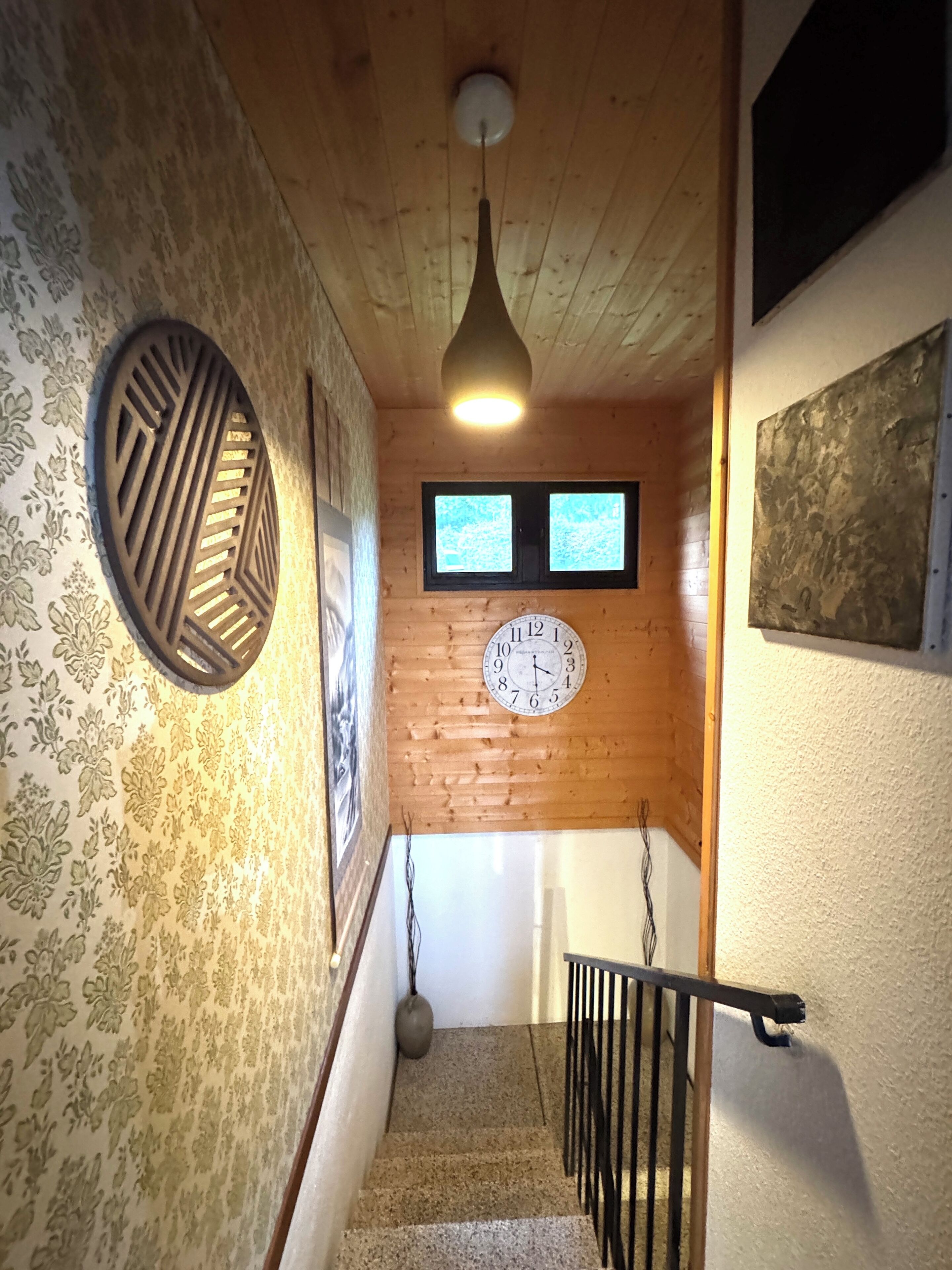 Un escalier en bois, une horloge, un papier peint à motifs et un luminaire suspendu.