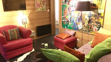En hyggelig stue med en rød sofa, en lille skammel og et fjernsyn på væggen.