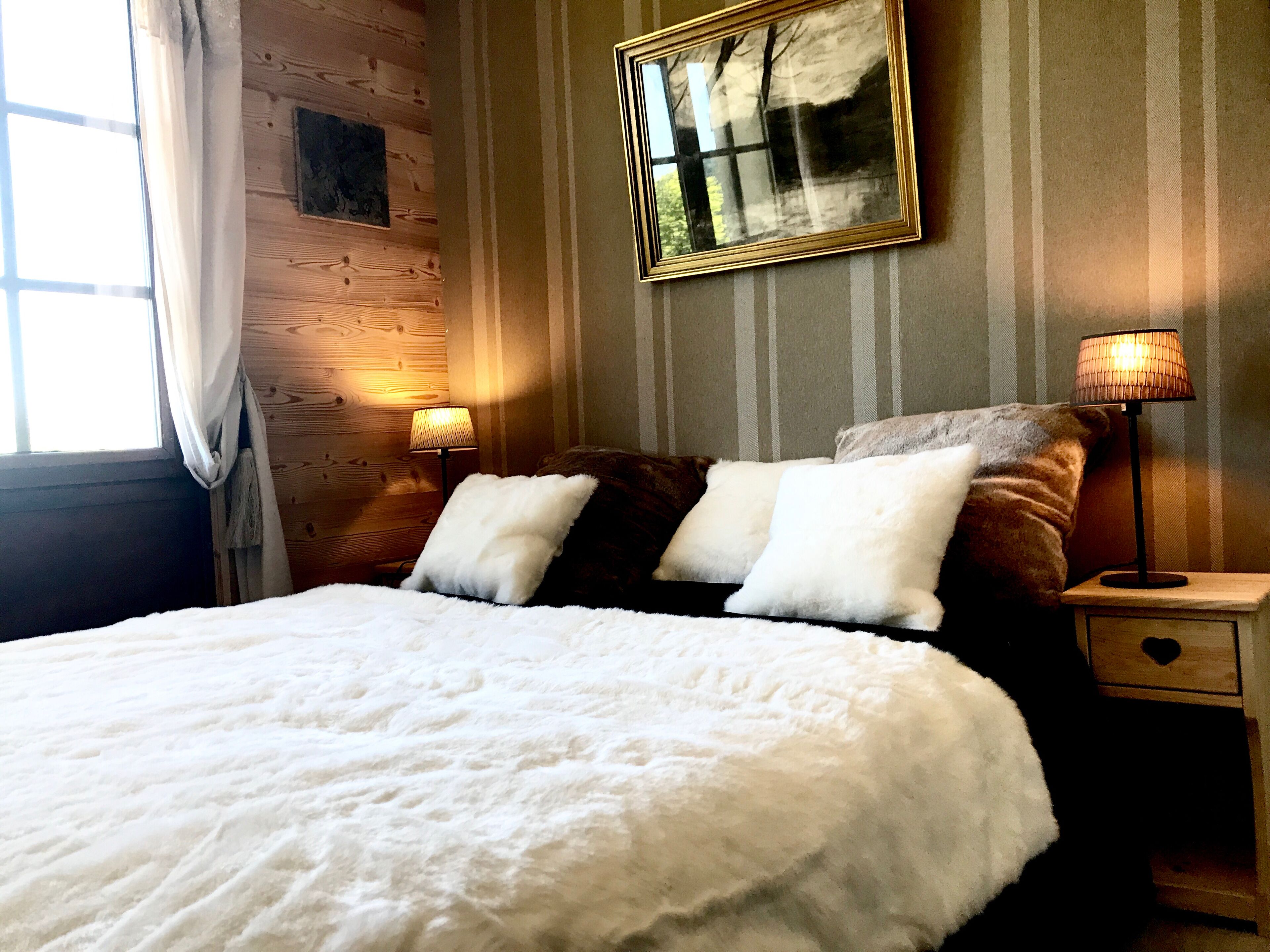 Een bed met wit beddengoed, een nachtkastje met een lamp, een ingelijst foto aan de muur en een raam met gordijnen.