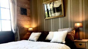 Una cama con ropa de cama blanca, una mesita de noche con lámpara, un cuadro en la pared y una ventana con cortinas.
