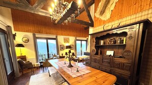 Una habitación acogedora con una mesa de comedor de madera, bancos y una araña. Hay sillas, una lámpara y vistas del exterior a través de amplias ventanas.