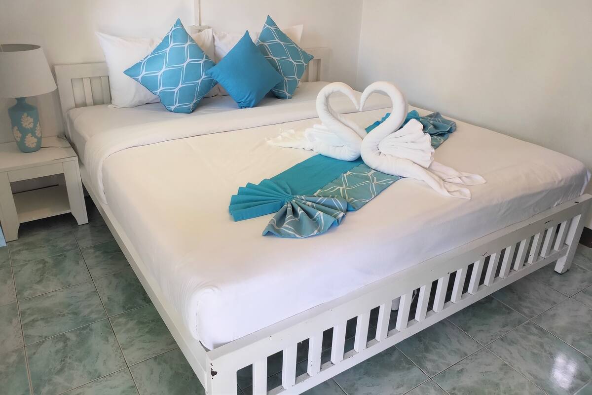 A neatly made bed with white bedding and blue pillows, a white nightstand with a lamp, and a decorative blue vase.