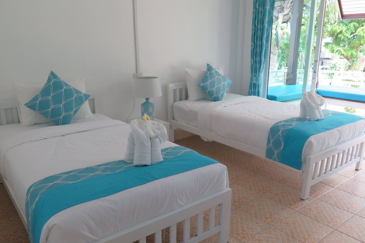 A hotel room with two beds, each with white bedding and blue accents, a small table with a lamp, and a view of the outdoors through a window with blue curtains.