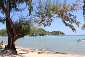A beach with a swing under a large tree, calm water, and boats in the distance. - Assava Dive Resort (Koh Tao)