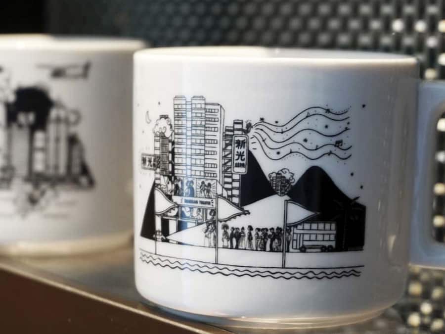 Two white mugs with black illustrations of urban scenes, including buildings and people.