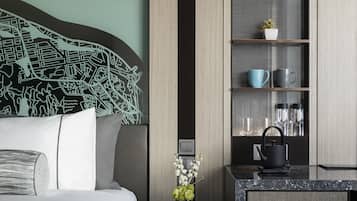 A modern hotel room with a bed, a nightstand with a vase of flowers, a coffee maker, and a shelf with decorative items.