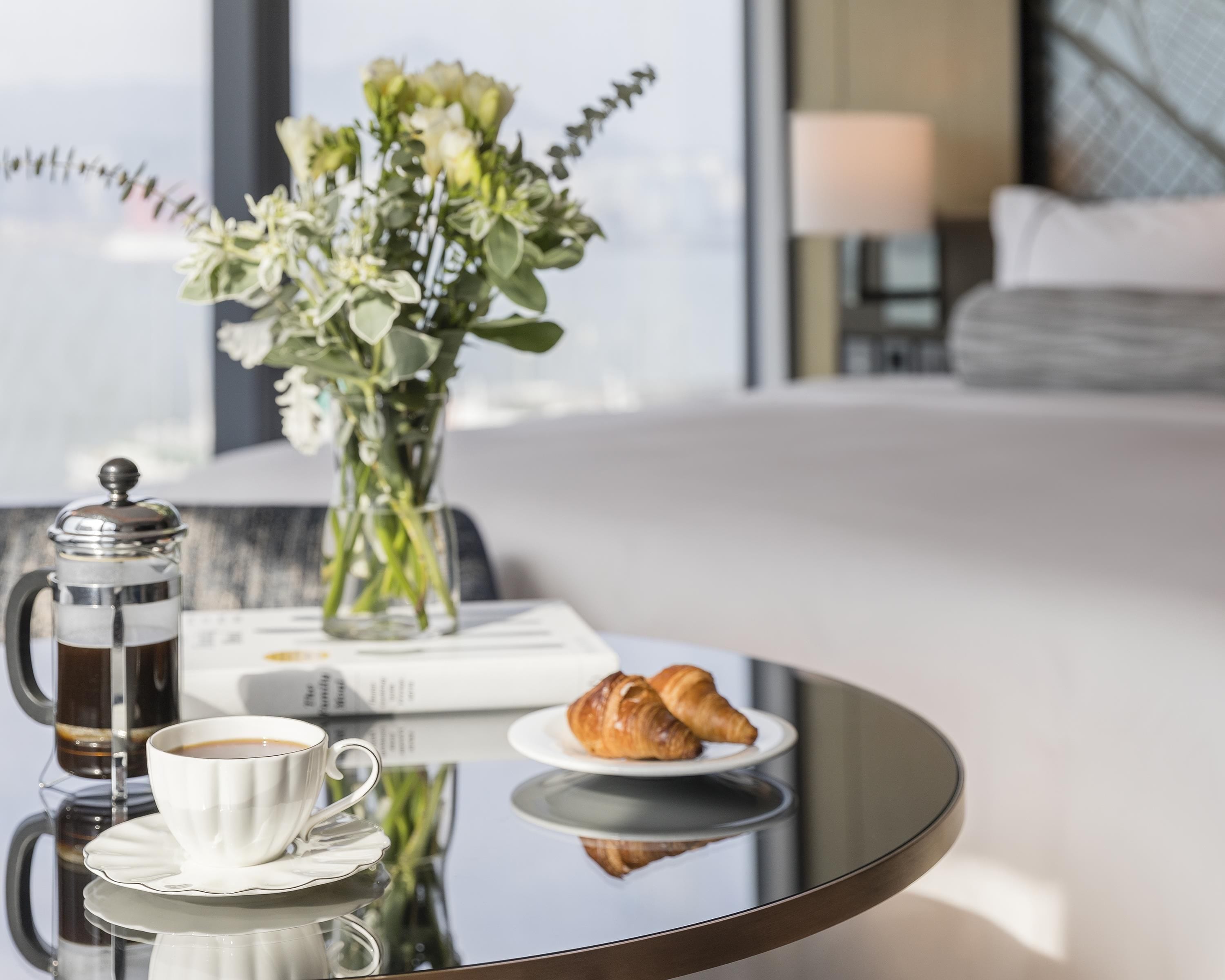 Quarto de hotel com uma mesa de centro de vidro, uma chávena de café branca, um prato com croissants e um vaso com flores.