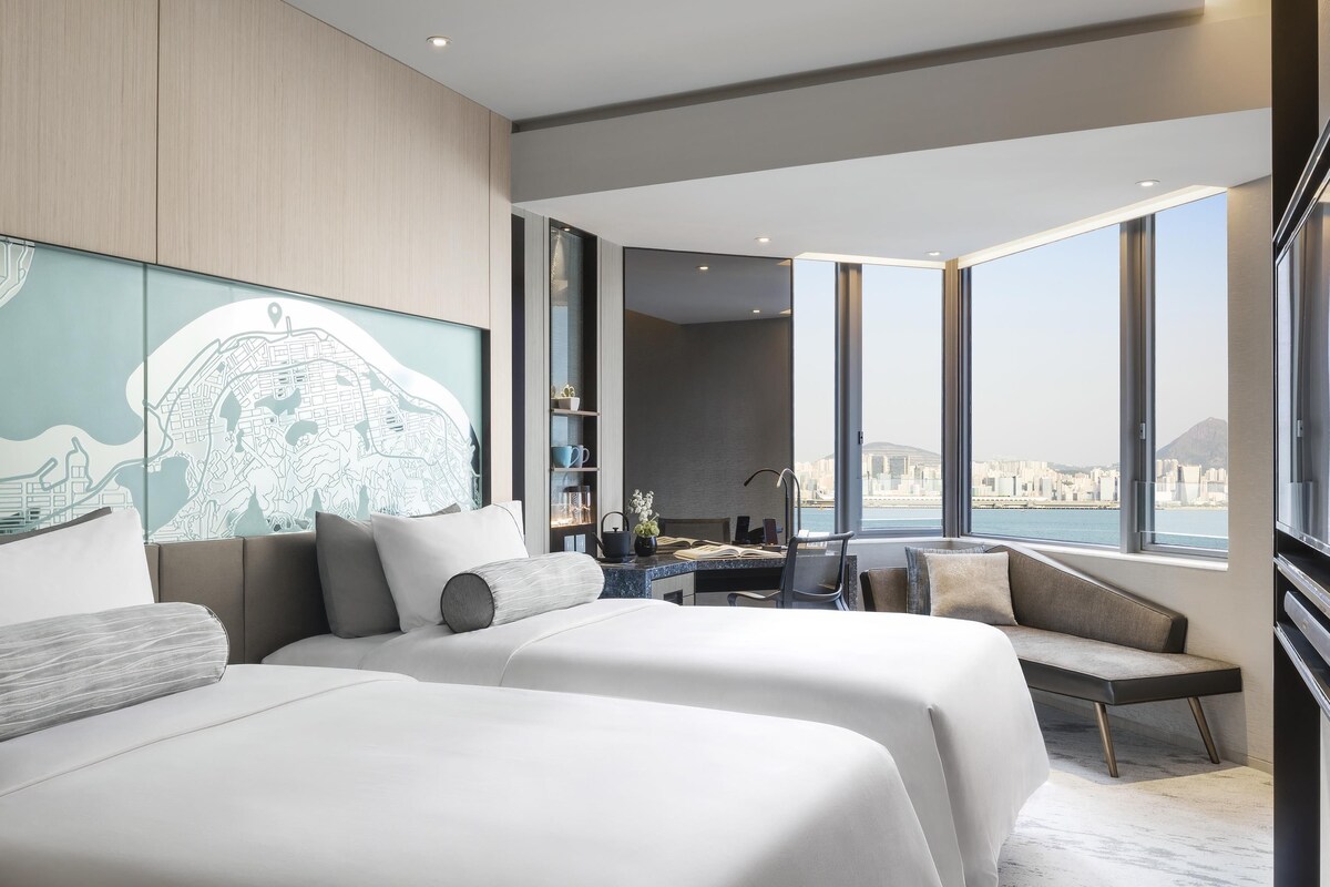 A modern hotel room with two beds, a desk, a chair, and a large window offering a city view.