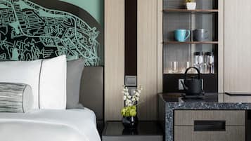 A modern hotel room with a neatly made bed, a small bedside table with a vase of flowers, a black kettle, and a built-in shelf with kitchen items.
