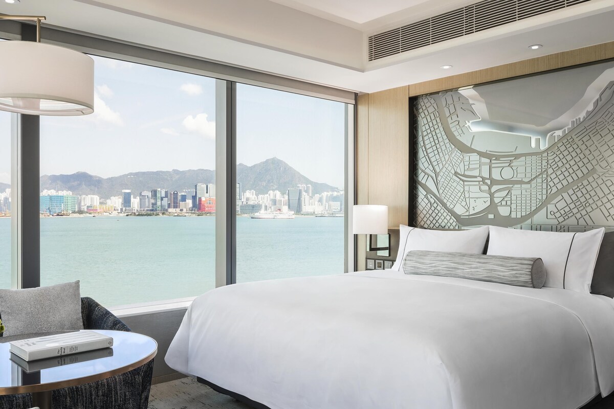 A hotel room with a large bed, a round table with a book, a chair, a city view, and a map wall.