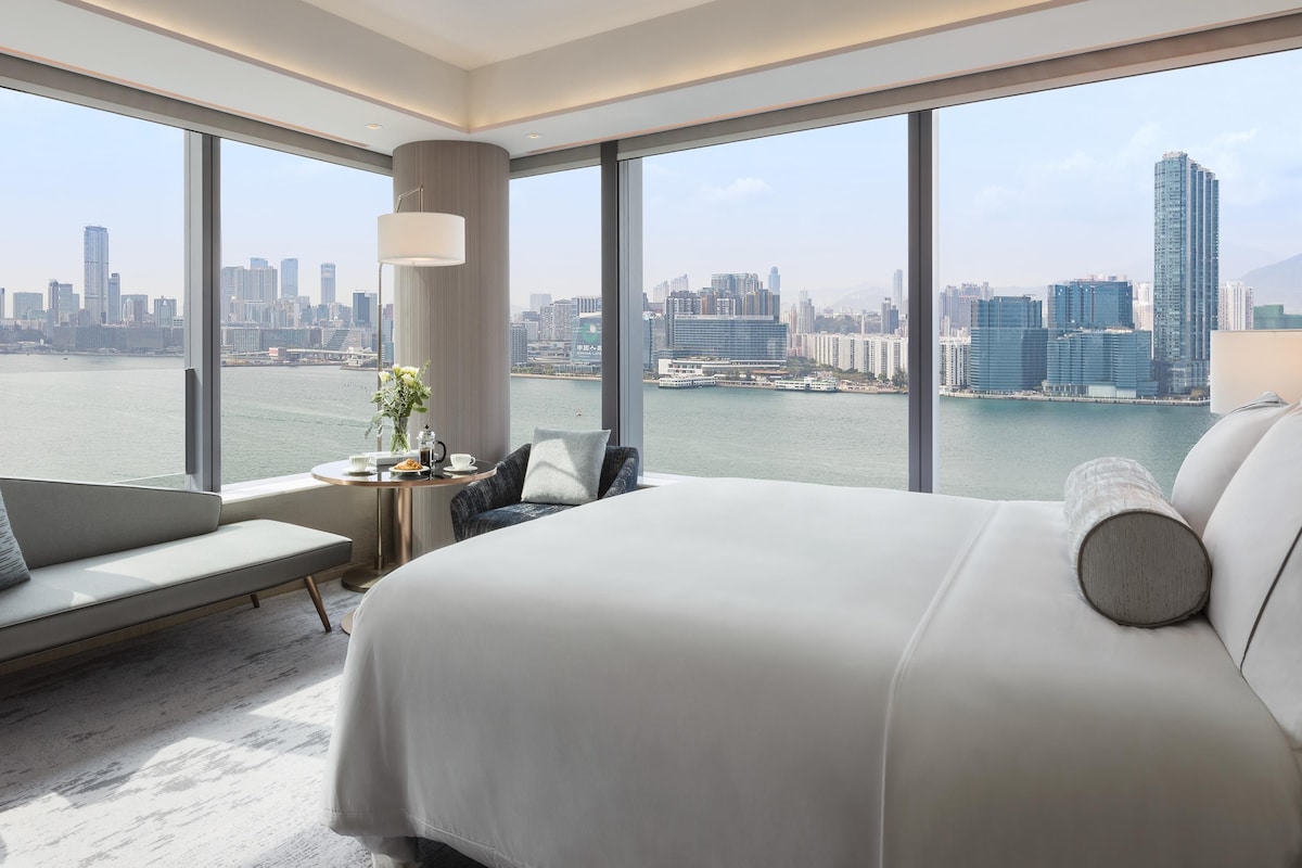 Una habitación de hotel moderna con una cama grande, una zona de estar y vistas a un horizonte urbano y al agua.