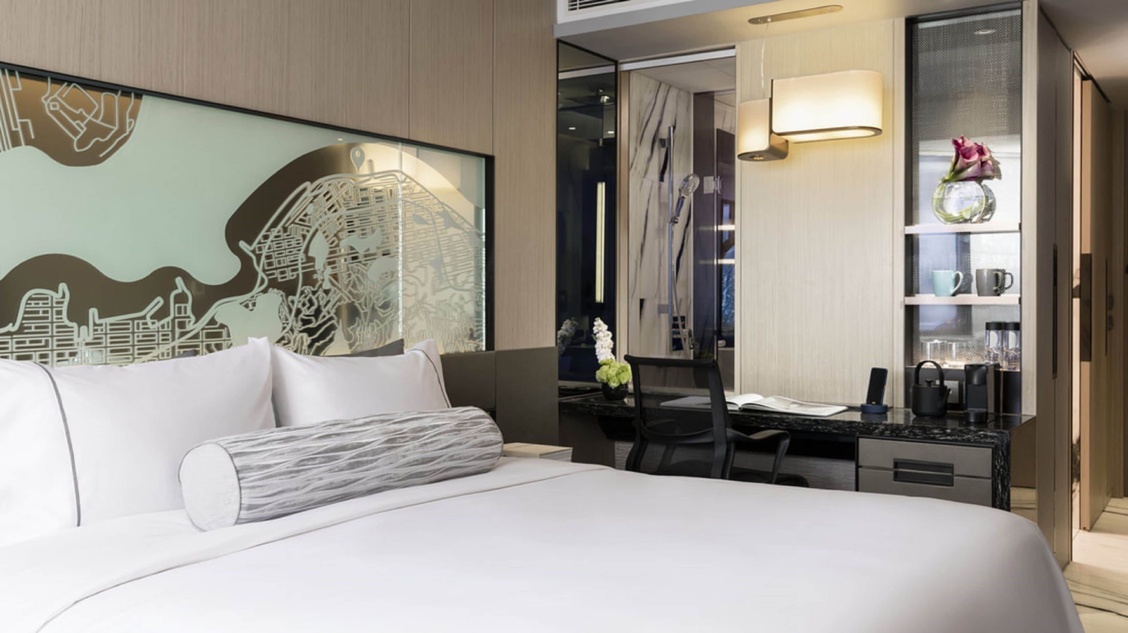 Une chambre d’hôtel moderne dotée d’un grand lit, d’un bureau avec un ordinateur, d’un petit coin cuisine et d’un mur décoratif orné d’un motif représentant un paysage urbain.