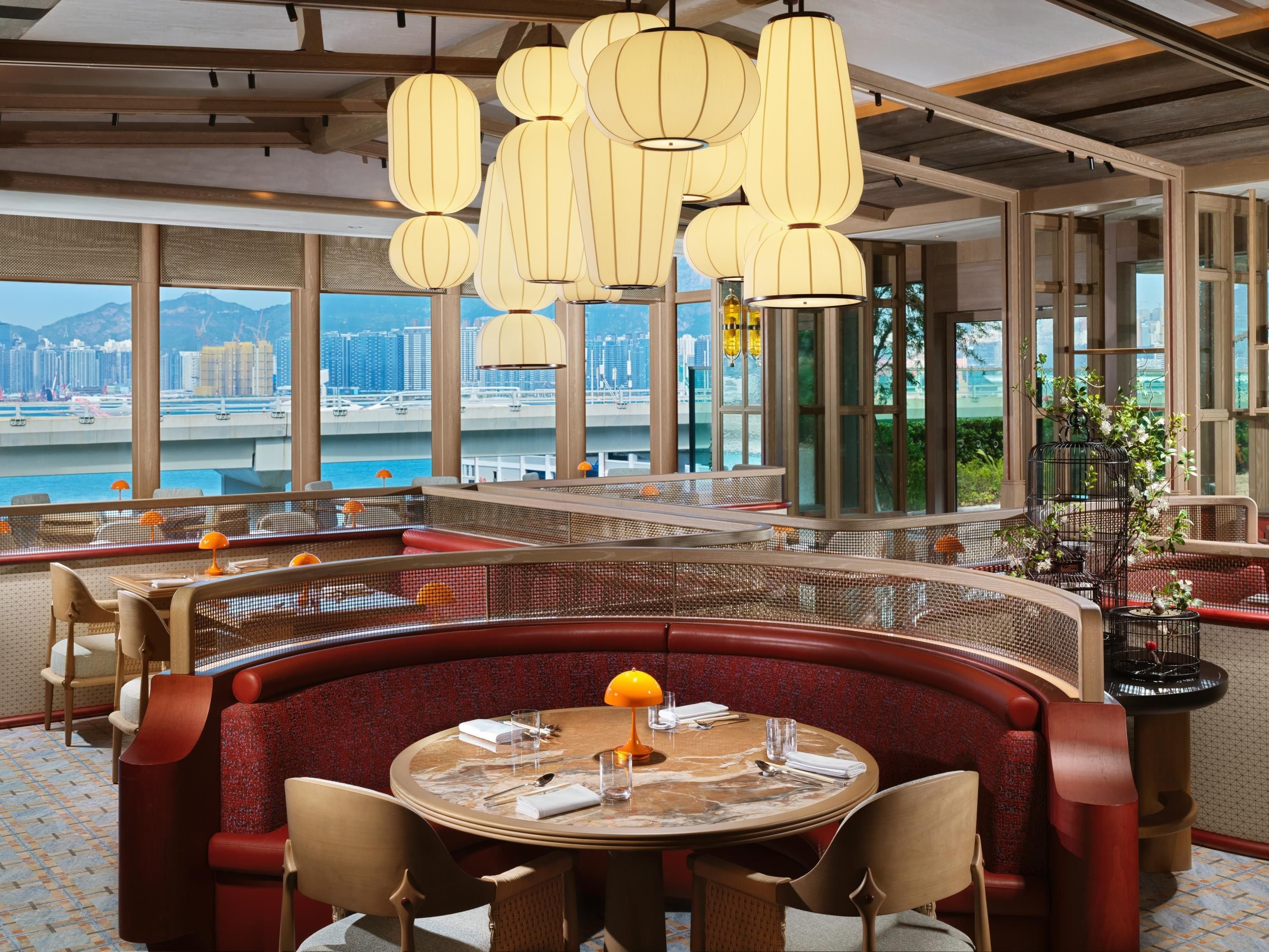 Un restaurante con mesas redondas, asientos tapizados en rojo y vistas a un paisaje urbano a través de grandes ventanales.