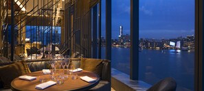 Un comedor con una mesa lista para cuatro personas, con vistas a un paisaje urbano y una masa de agua al anochecer.