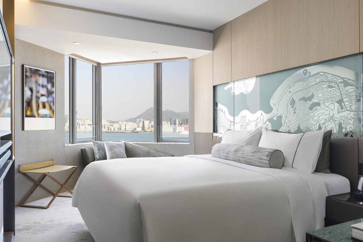 A modern hotel room with a large bed, a desk, and a view of a cityscape through large windows.