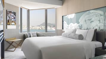 A modern hotel room with a large bed, a desk, and a view of a cityscape through large windows.