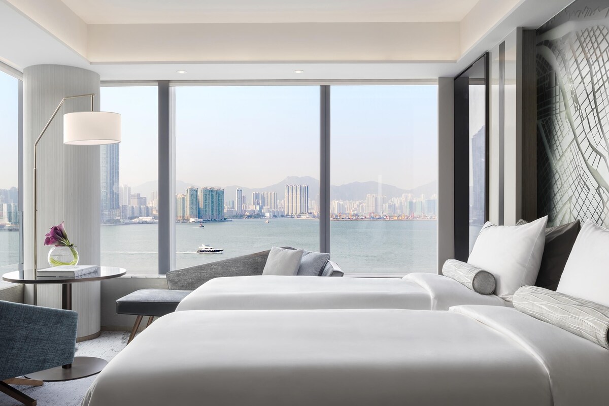 A modern hotel room with a large window, a bed, a sofa, and a desk with a lamp.