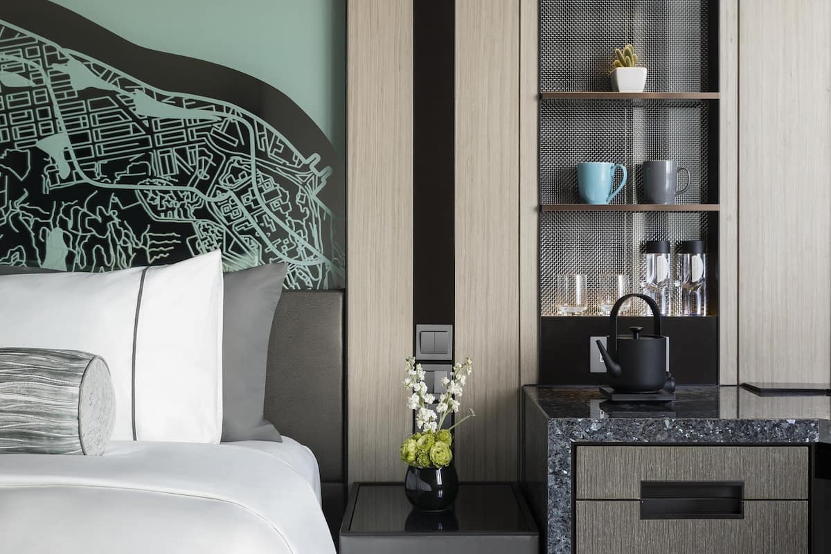 Habitació d'hotel moderna amb un llit, una tauleta de nit amb un ram de flors, una cafetera i una estant amb objectes decoratius.