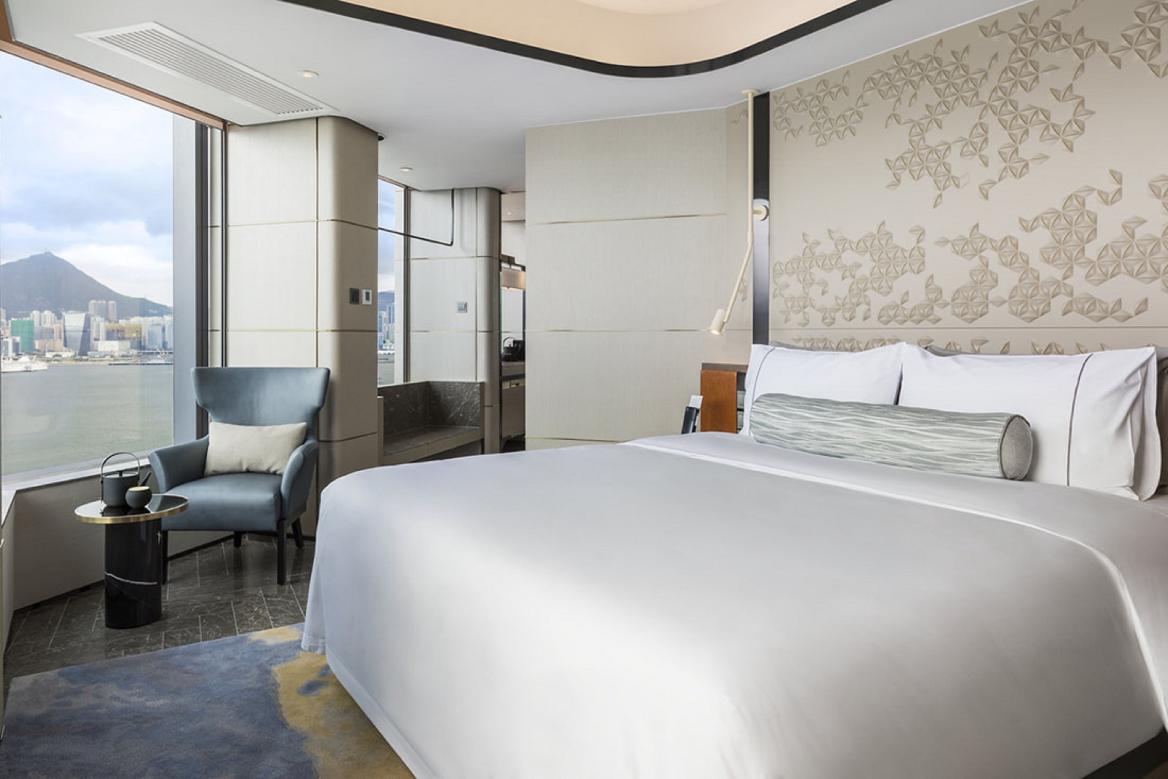 A modern hotel room with a large bed, a seating area with a chair, a small table, and a view of a cityscape through large windows.