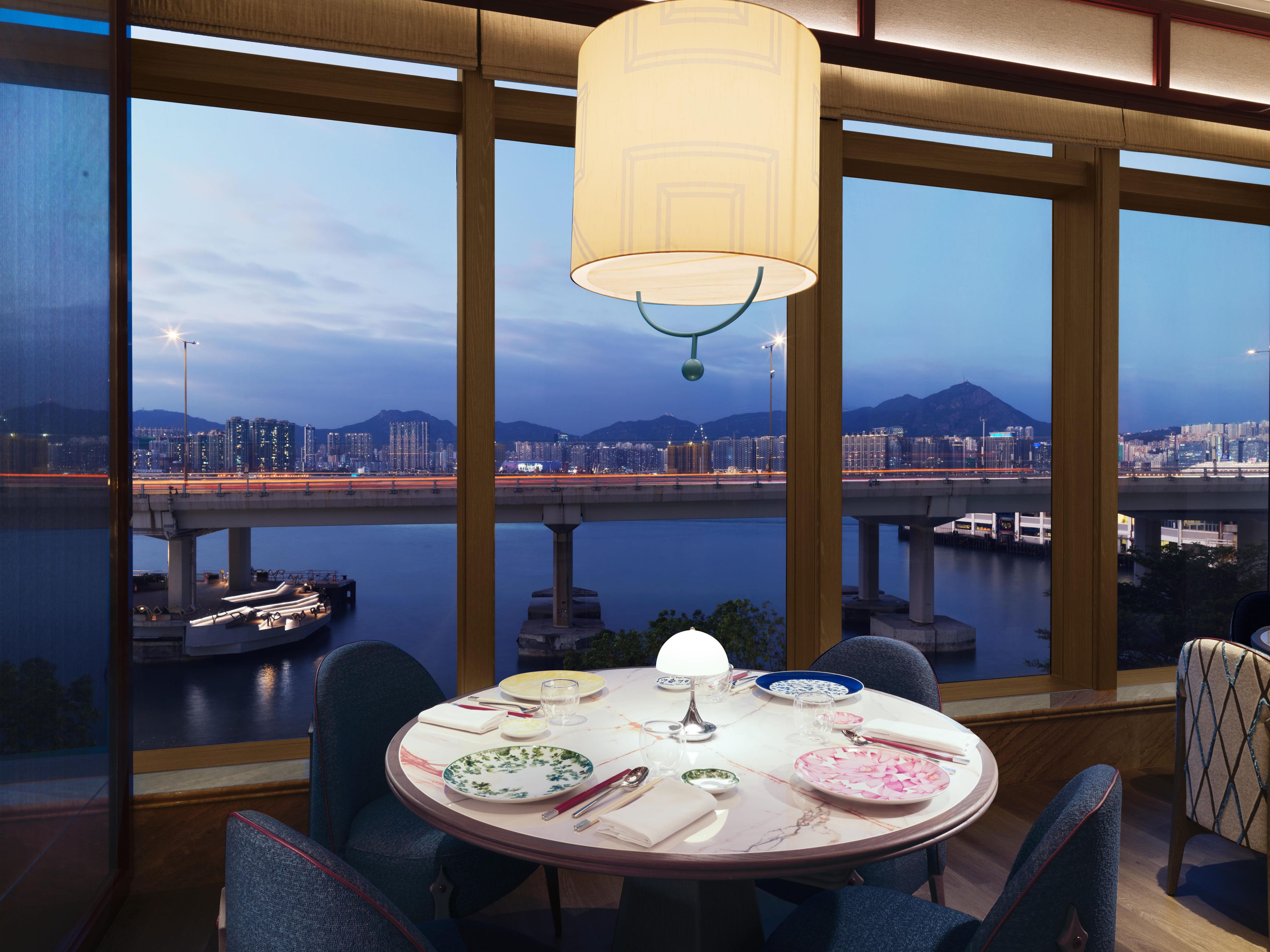 Un comedor con una mesa dispuesta para cuatro personas, ventanas amplias con vistas a la ciudad y una lámpara colgante.