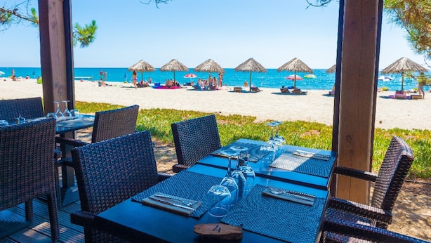 An outdoor dining area with tables and chairs, overlooking a beach with umbrellas and people. - Standing Apartment, Facing the Sea! (Ghisonaccia)