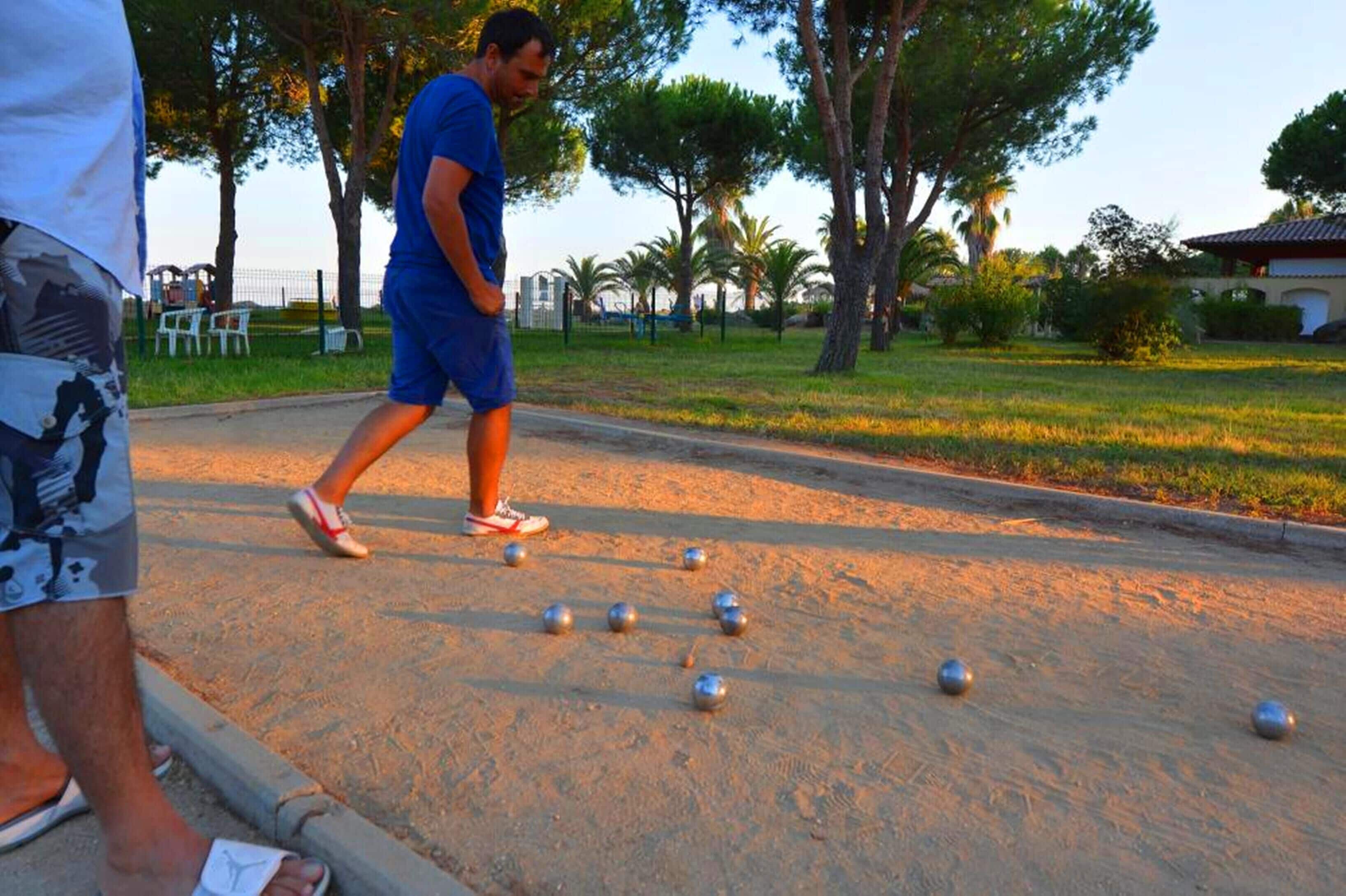 A person playing bocce on a sandy court with trees and a building in the background.