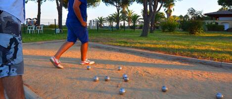 A person playing bocce on a sandy court with trees and a building in the background.