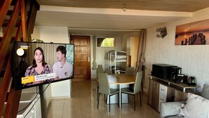 A living room with a TV displaying a news program, a dining area with a table and chairs, and a kitchen area with a microwave and coffee maker. - Standing Apartment, Facing the Sea! (Ghisonaccia)