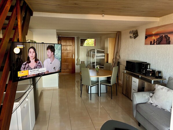 A living room with a TV displaying a news program, a dining area with a table and chairs, and a kitchen area with a microwave and coffee maker. - Standing Apartment, Facing the Sea! (Ghisonaccia)