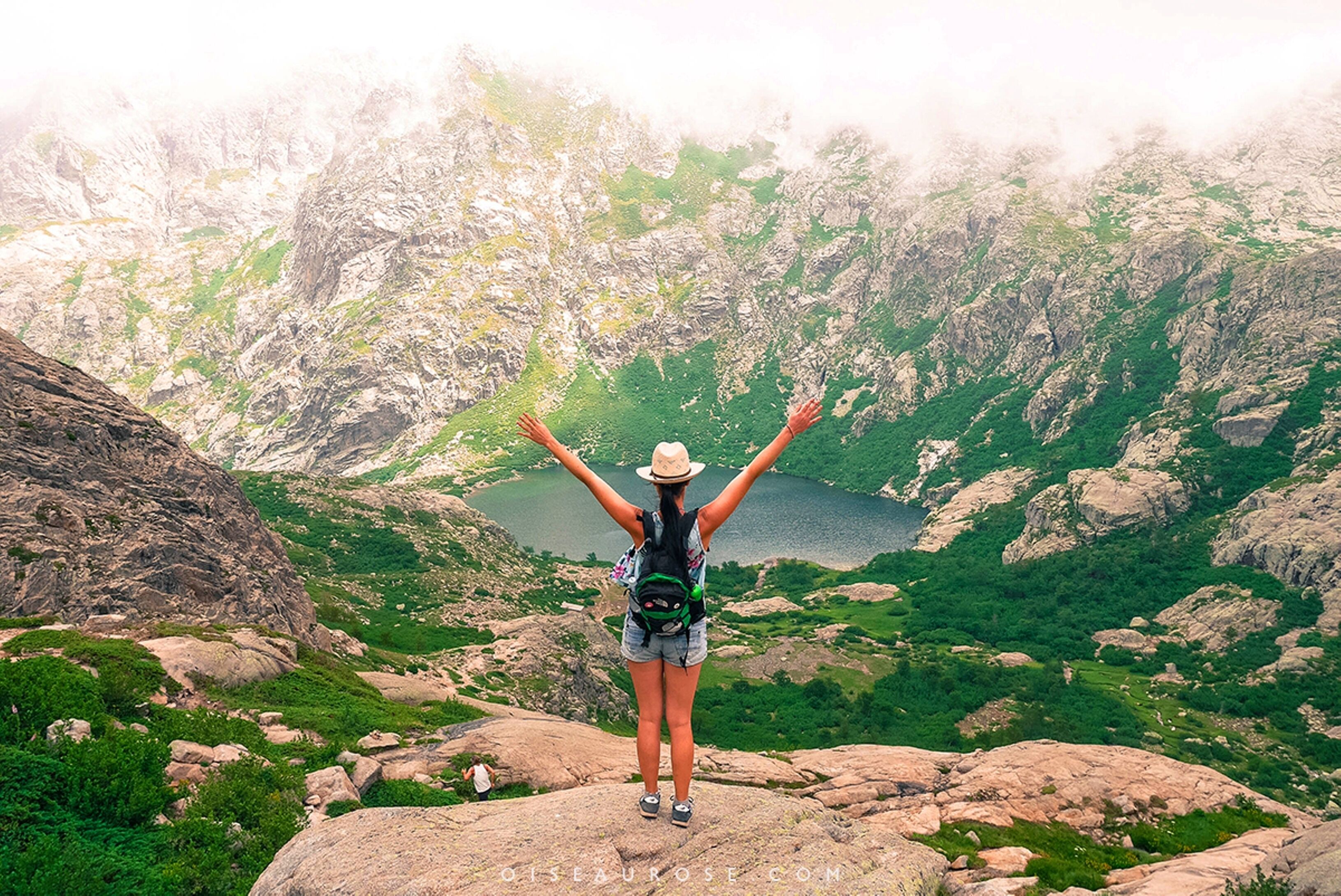 Une personne se tient debout sur un promontoire rocheux, les bras levés, et surplombe un paysage montagneux avec un lac.