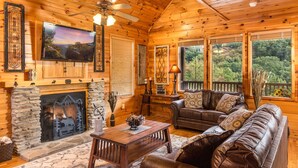 A cozy log cabin interior with a fireplace, leather sofa, and large windows offering a view of the forest.