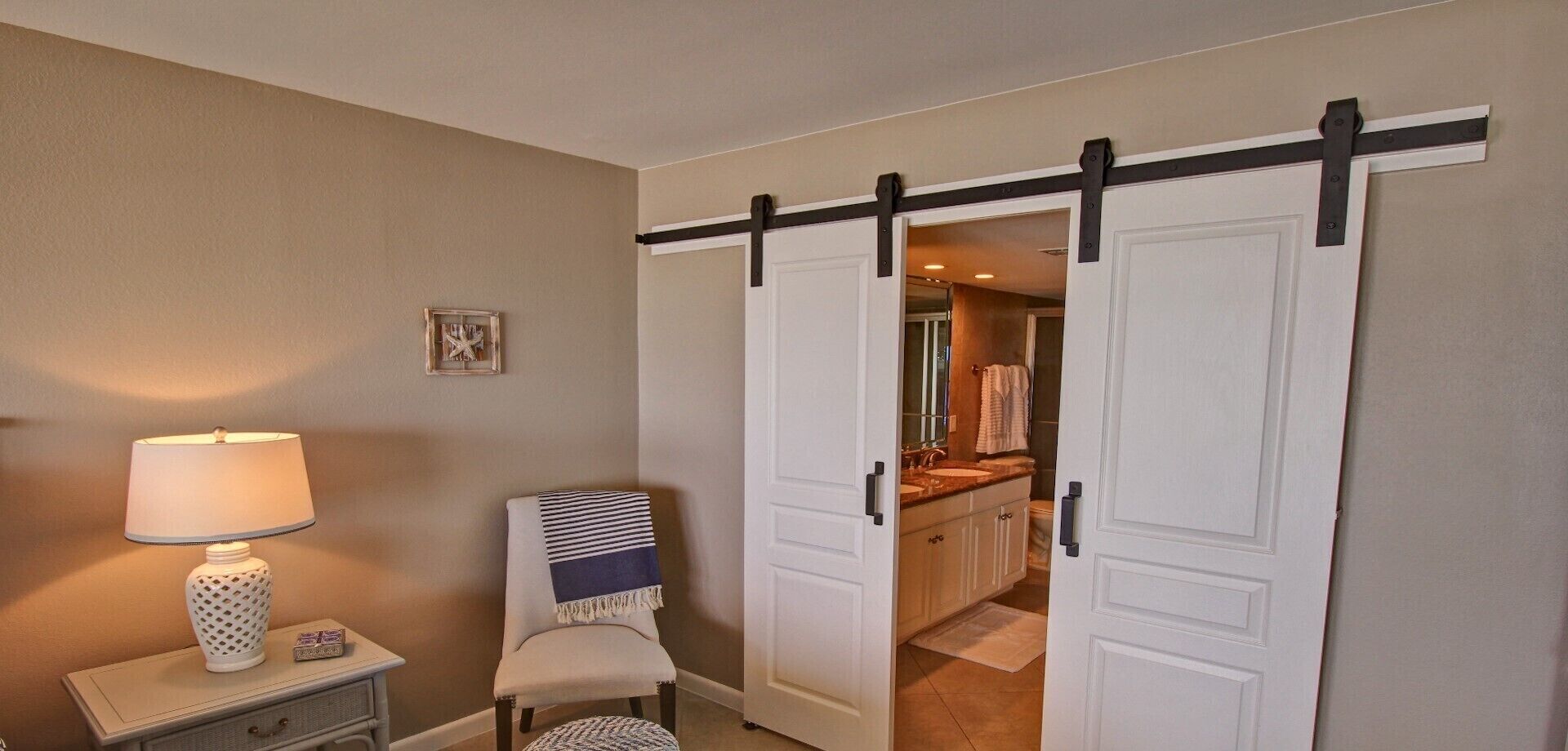 A room with a sliding door leading to a bathroom, a bedside table with a lamp, and a chair with a striped blanket.