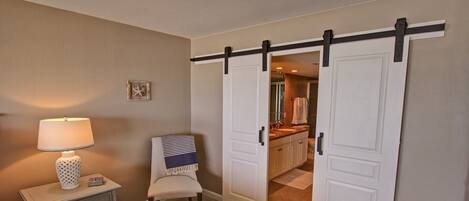 A room with a sliding door leading to a bathroom, a bedside table with a lamp, and a chair with a striped blanket.