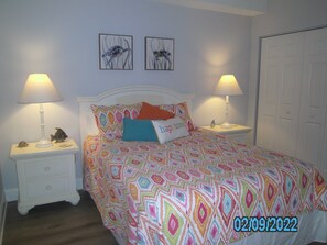 Ein Schlafzimmer mit einem geblümten Bettüberwurf, zwei an der Wand befestigten Bildern, zwei Nachttischlampen und einer weißen Tür.