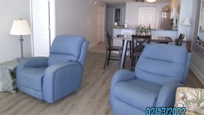 Un salon avec deux fauteuils bleus, une table à manger avec des chaises, un coin cuisine et un lustre.