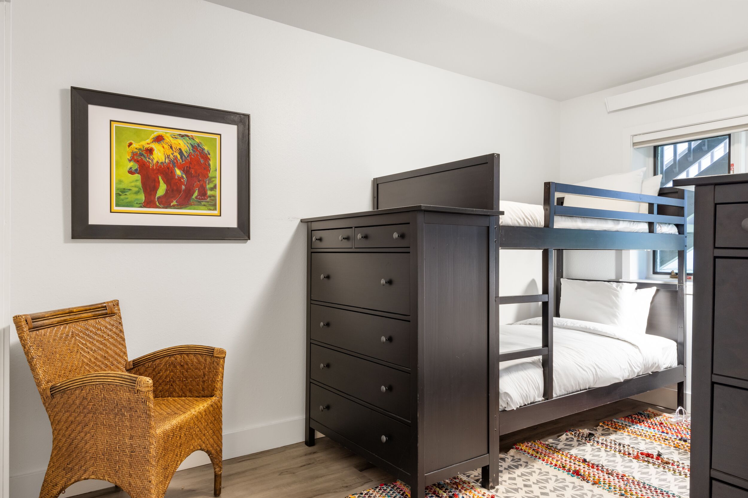 A bedroom with a bunk bed, a dresser, a wicker chair, and a framed picture on the wall.
