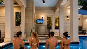 Un área de piscina con cuatro personas sentadas en plataformas flotantes, una terraza cubierta y un televisor montado en la pared.