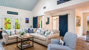 Amplia sala de estar con techo alto, ventana grande y un cartel que dice 'BEACH HOUSE'.