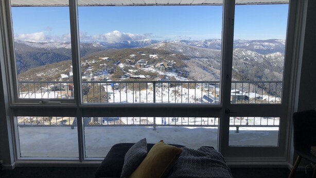 A room with a large window overlooking a snowy mountain landscape, a couch, and a wooden table. - Twin Towers 701A (Mount Buller)