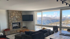 A modern living room with a stone fireplace, a large TV, and a dining area with a view of mountains. - Twin Towers 701A (Mount Buller)