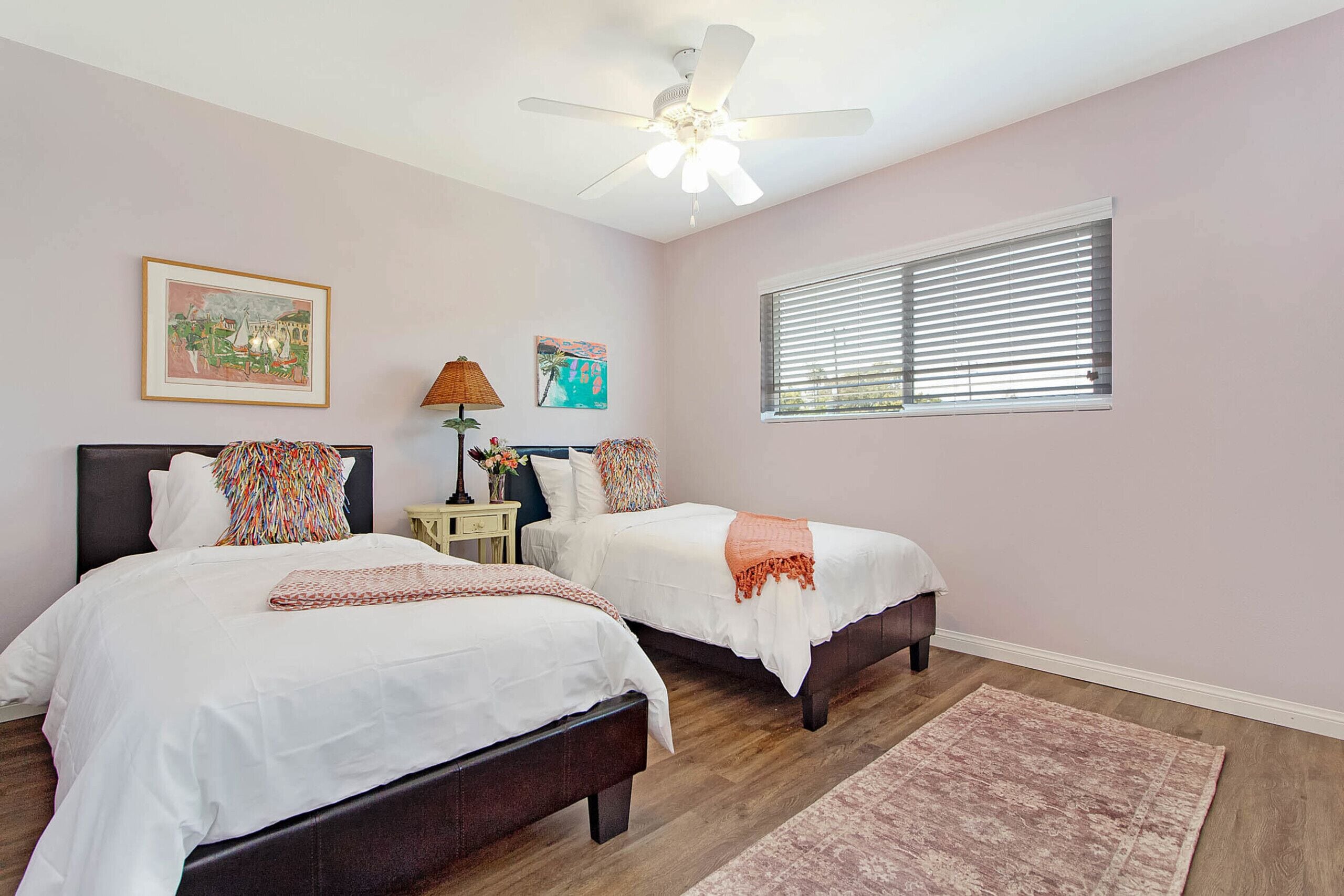 A bedroom with two beds, a ceiling fan, a window with blinds, and artwork on the walls.