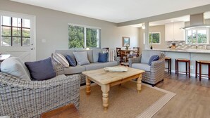 A modern living room with a sofa, coffee table, wicker chairs, and a kitchen area with bar stools.