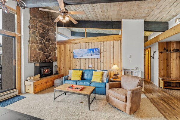 Ein gemütliches Wohnzimmer mit Steinwand, Holzbalkendecke, einem blauen Sofa, einem braunen Sessel und einem Kamin.