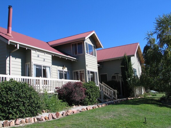 A two-story house with a red roof, a front porch with a white railing, and a stone retaining wall. - Alpine Stag Lodge House (Merrijig)