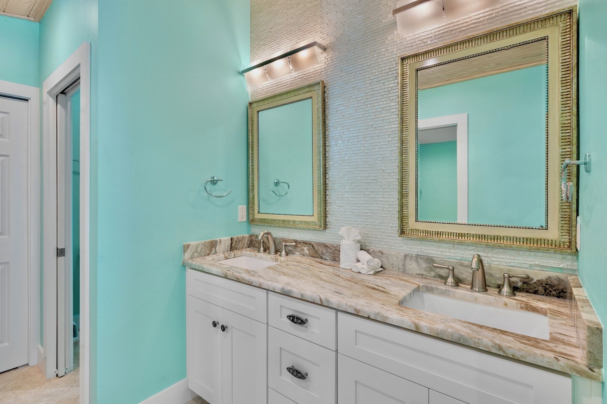 A bathroom with a turquoise wall, two large mirrors with gold frames, a marble sink, and a white cabinet with drawers.